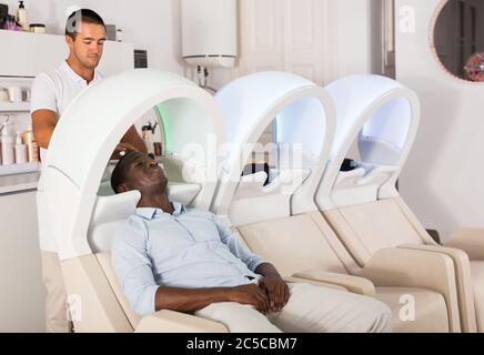 Ein frohes afroamerikanischer Mann, der sich am Waschbecken anlehnte und sich beim Friseursalon die Haare wusch Stockfoto