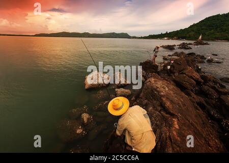 Zwei asiatische Fischer fischen in der Dämmerung vom Felsen an einer Küste, dramatische Wolken und der Sonnenuntergang Himmel im Hintergrund. Konzentrieren Sie sich auf den Fischer. Stockfoto