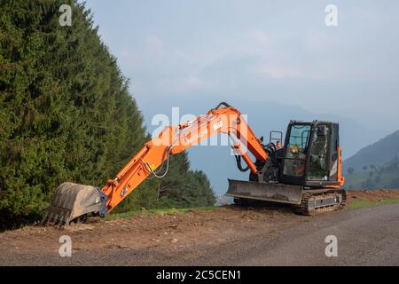 Hitachi Raupenbagger geparkt auf einer neu gebauten Hochgebirgsstraße vor einem grünen Nadelwald im Hintergrund, Lombardei, Italien Stockfoto