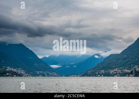 Panoramablick auf den Comer See an einem regnerischen Sommertag, italienische Alpen und stürmischer Himmel im Hintergrund. Blick von der Küstenpromenade der Stadt Como. Stockfoto
