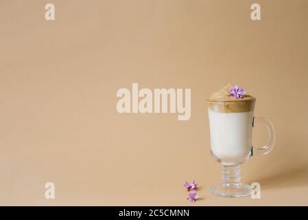 Schöner Dalgon-Kaffeedrink mit Schaum in einer transparenten Tasse und lila Blüten auf beigem Hintergrund. Speicherplatz kopieren Stockfoto