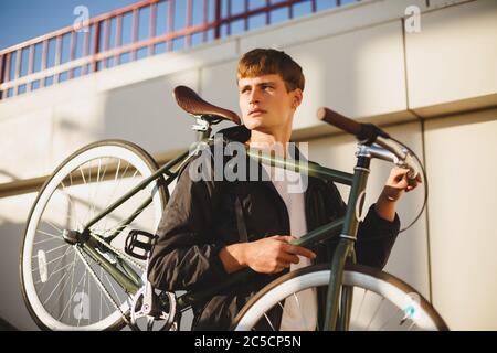 Nachdenklicher Junge mit braunen Haaren stehend und Fahrrad haltend, während er traumhaft zur Seite schaut. Junger Mann in Daunenjacke stehend mit klassisch schön Stockfoto