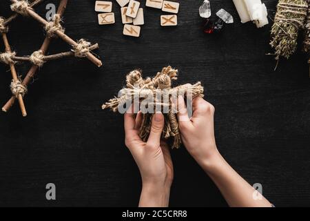 Draufsicht auf Frau, die Voodoo-Puppen in der Nähe skandinavischer Runen, Pentagramm, Kerzen und Kristalle auf Schwarz hält Stockfoto