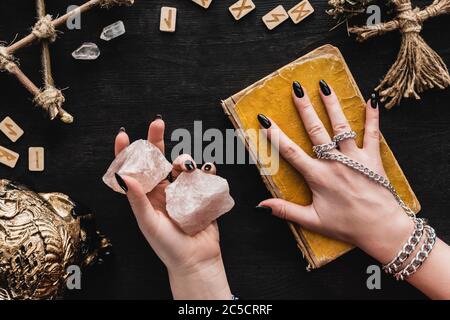 Draufsicht auf Hexe mit Kristallen und berührenden gealterten Buch der dunklen Magie in der Nähe Voodoo Puppe und Runen auf schwarz Stockfoto