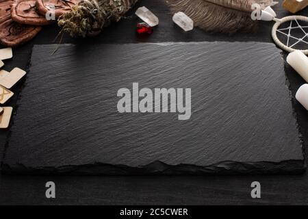 Schwarzes Brett neben Holzrunen, Kerzen und Traumfänger auf schwarzer Holzfläche Stockfoto