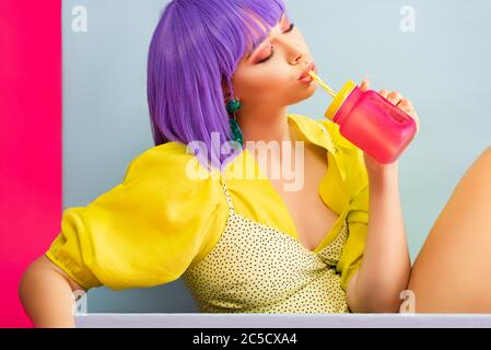 Schöne Pop Art Mädchen in lila Perücke als Puppe trinken aus Glas, während in blauen Box sitzen, isoliert auf rosa Stockfoto