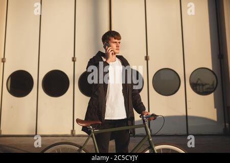 Junger Mann mit braunen Haaren steht mit klassischem Fahrrad und sieht beim Telefonieren auf seinem Handy traumhaft zur Seite. Durchdachte junge in Daunenjacke Stockfoto