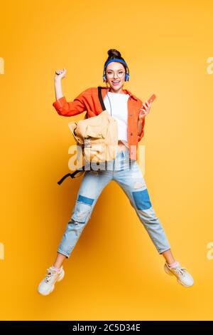 Fröhliche Schüler in Headset und Brille springen, während das Smartphone auf gelbem Hintergrund halten Stockfoto