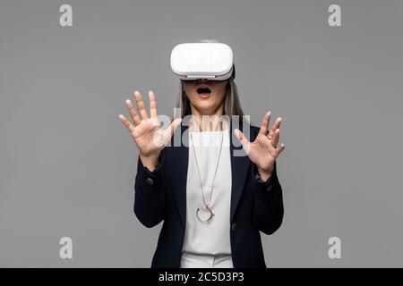 Schockiert Geschäftsfrau gestikulieren und mit Virtual-Reality-Headset isoliert auf grau Stockfoto