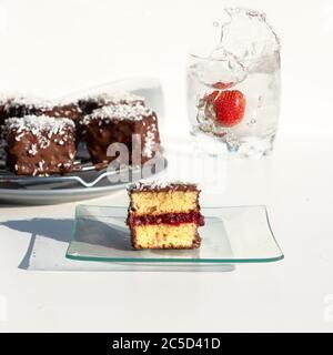 Traditionelle australische lamington Kuchen mit Erdbeermarmelade, Schokolade und Kokosnuss. Gegen ein Glas Wasser, ein Spritzer Wasser, fallen Stockfoto