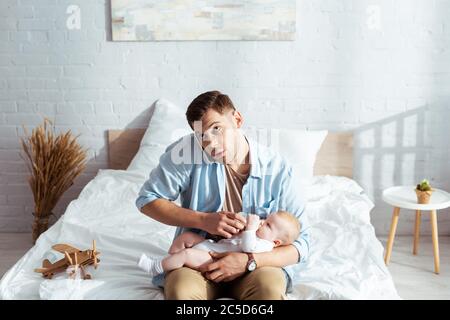 Aufmerksamer Mann, der auf dem Smartphone spricht, während er niedlichen kleinen Sohn aus der Babyflasche füttert Stockfoto