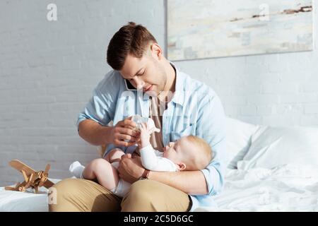 Junger Vater füttert Baby Junge aus Babyflasche während des Sprechens auf dem Smartphone Stockfoto