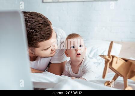 Selektiver Fokus des jungen Vaters, der überrascht Baby beim Liegen im Bett in der Nähe von Laptop und Spielzeug Flugzeug Stockfoto