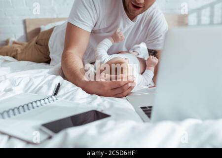 Beschnittene Ansicht des Mannes, der mit entzückenden Kind in der Nähe von Gadgets und Notebook auf dem Bett spielt Stockfoto