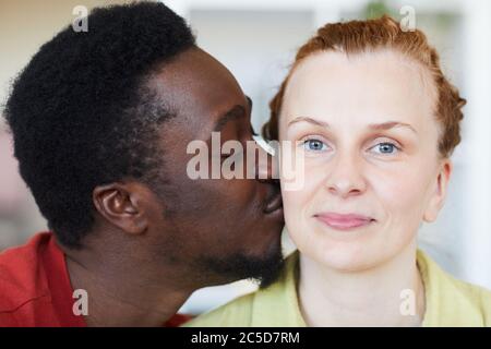Nahaufnahme einer jungen Frau, die die Kamera anschaut, während ein afrikanischer Mann ihre Wange küsst Stockfoto