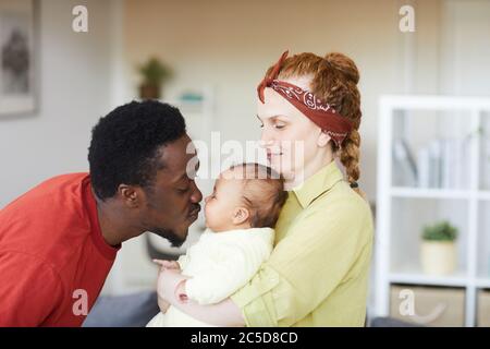 Junge Frau hält Baby an den Händen, während afrikanischer Mann sie küsst, sind sie zu Hause Stockfoto
