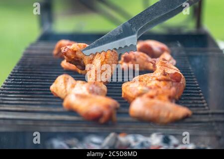 Gegrillte Hähnchenflügel auf dem Grill mit Rauch, Metallzangen zu drehenden Hähnchenflügel. Nahaufnahme, selektiver Fokus Stockfoto