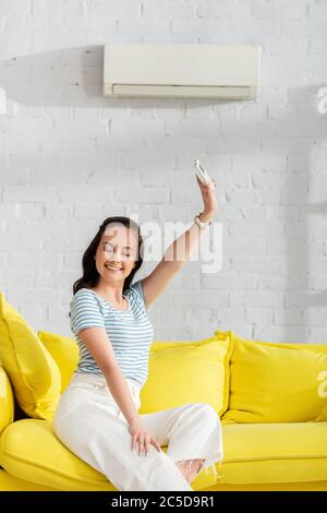 Lächelnde Frau mit Fernbedienung der Klimaanlage auf dem Sofa im Wohnzimmer Stockfoto