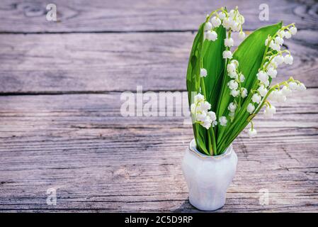 Lilien des Tales schöne Frühlingsblumen stehen Bouquet in einer Vase auf einem alten Holztisch. Maiglöckchen. Blume Frühling Sonne Weiß Grün Rückseite Stockfoto