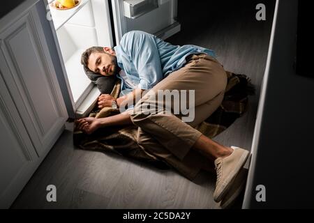 Schöner Mann, der nachts in der Küche neben einem offenen Kühlschrank schläft Stockfoto