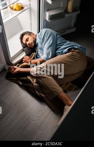 Man konnte aus der Perspektive sehen, wie der Mann nachts in der Nähe eines offenen Kühlschranks auf dem Boden schlief Stockfoto