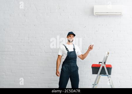 Ein hübscher Arbeiter in den Overalls lächelte und hielt die Fernbedienung der Klimaanlage neben Werkzeugkasten und Leiter Stockfoto