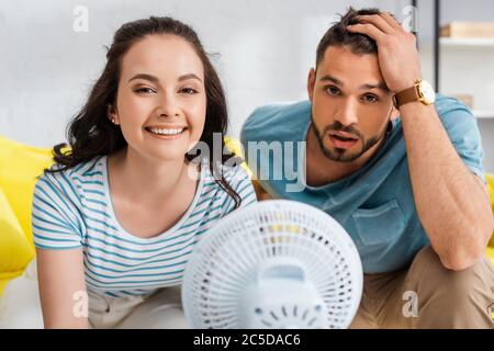 Selektive Fokus der lächelnden Mädchen Blick auf die Kamera in der Nähe müde Freund und elektrischen Ventilator Stockfoto
