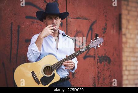 Der gutaussehende Mann trägt einen Cowboy-Hut, raucht eine Zigarette und hält eine akustische Gitarre Stockfoto