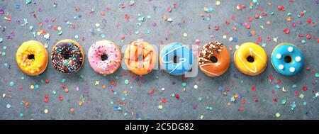 Verschiedene bunte Donuts in einer Reihe mit Konfetti auf rustikal grauem Hintergrund Stockfoto