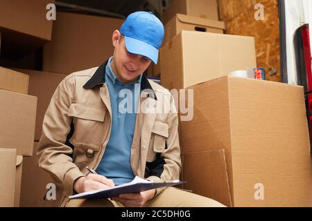 Der Zusteller füllt die Dokumente, während er draußen mit Kartons im Hintergrund sitzt Stockfoto