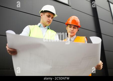 Mann und Frau in Arbeit Helme halten Blaupause des neuen Gebäudes und untersuchen es im Team, während im Freien stehen Stockfoto