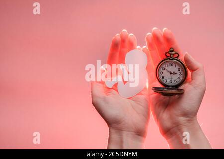 Papier Embryo Silhouette und Uhr in Frauenhand mit Licht. Heller korallenfarbener Hintergrund mit Kopierbereich. Hände befinden sich auf der rechten Seite. Weichfokus. Stockfoto