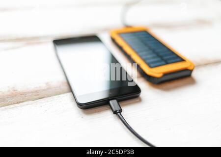 Konzept moderner Technologien. Smartphone mit schwarzem Bildschirm, aufgeladen von einem tragbaren Solarladegerät. Weißer Holzhintergrund mit Streifen. Nahaufnahme. Stockfoto