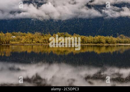 Erstaunliche Herbstansicht mit flauschigen Wolken und Nebel über Bäumen, See und Bergen nach Regen und ihre Reflexion im glatten Wasser des Sees Stockfoto