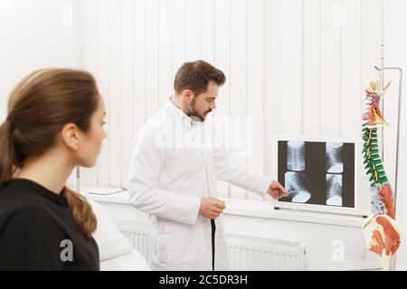 Der Arzt zeigt dem Patienten Ergebnisse der Röntgenuntersuchung und erklärt die Ursache ihrer Schmerzen in der Arztpraxis. Patient besucht Physiotherapeut in Stockfoto