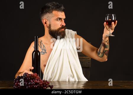 Sommelier untersucht Getränk. Weinbau und Weinlese Konzept. Gott Bacchus mit aufmerksamem Gesicht trägt weiße Tuch sitzt von Flasche und Trauben. Mann mit Bart hält ein Glas Wein auf braunem Hintergrund Stockfoto