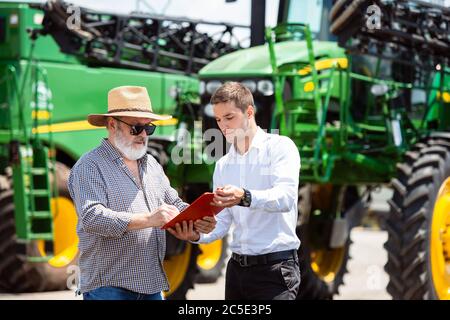 Vereinbarung. Professioneller Landwirt mit einem modernen kombinieren in Sonnenlicht bei der Arbeit. Selbstbewusste, helle Sommerfarben. Landwirtschaft, Ausstellung, Maschinen, Pflanzenproduktion. Senior Mann in der Nähe seines Traktors mit Investor. Stockfoto