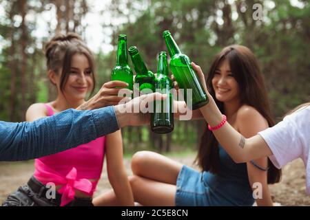 Glück. Gruppe von Freunden klirrende Bierflaschen während Picknick im Sommerwald. Lifestyle, Freundschaft, Spaß haben, Wochenende und Ruhe Konzept. Sieht fröhlich, glücklich, feiern, festlich. Stockfoto