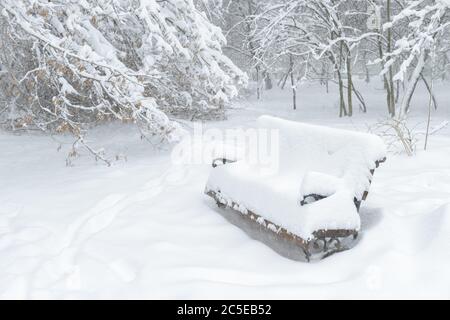 Schneebank während Schneesturm im Park, Moskau, Russland. Winter Park Hintergrund. Verschneite Bank auf der Straße. Bank in einer Schneewehe. Schneefall im Stockfoto