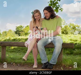 Junge Liebhaber, die an einem sonnigen Sommertag im Springfield Park auf der Bank im Park auf das Mobiltelefon schauen. Model Release verfügbar Stockfoto