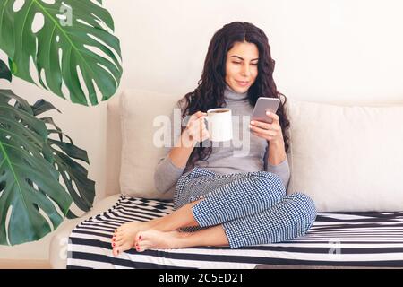 Kaukasische Frau mit Smartphone, während entspannt trinkt Morgenkaffee sitzen bequem auf der Couch, in sozialen Netzwerken surfen und lesen Sie die Nachrichten. Stockfoto