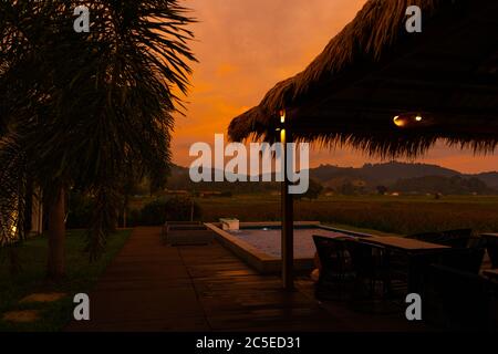 Schöner orange feuriger Sonnenuntergang in den Tropen. Straßendach von Heu, neben dem Pool und mit Blick auf das Reisfeld Stockfoto