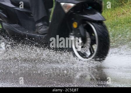 Motorrad Moped fährt durch eine Pfütze auf einer nassen Straße im Regen. Spray fliegt von den Rädern. Nahaufnahme Stockfoto