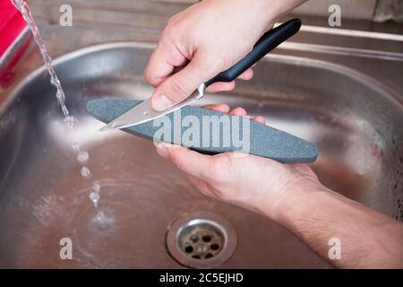 Nahaufnahme Hände eines Mannes sorgfältig manuell schärfen ein Messer unter einem Strom von Wasser auf einem Schleifstein. Schärfen für den Haushalt. Pflege für Küchenbesteck. Stockfoto