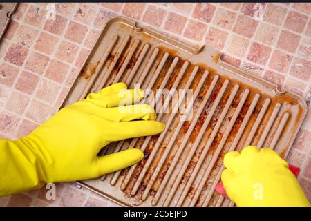 Hände in gelben Haushaltshandschuhen entfernen Fett von einem schmutzigen elektrischen Grill mit einem Schwamm und einem speziellen Spray. Tropfen Fett nach dem Braten. Stockfoto