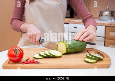 Nahaufnahme der Hände einer jungen Frau, die mit einem Messer in Scheiben oder Scheiben von jungen Zucchini-Gurken auf einem hölzernen Schneidebrett geschnitten wird. Zubereitung der Zutat Stockfoto