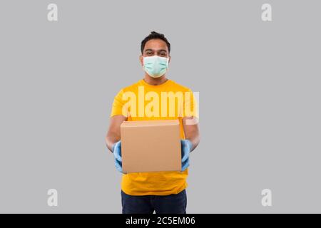 Indian man mit Box in den Händen Tragen medizinische Maske und Handschuhe isoliert. Gelbe T-Shirt Lieferung Junge. Lieferung Nach Hause. Quarantäne-Held. Mann Lächelt Stockfoto