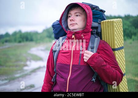 Guy Tourist Wanderer mit Rucksack und Reisematte, in einer roten Jacke steht im Regen Stockfoto