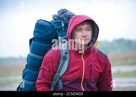 Ein Kerl mit Rucksack steht in der Nähe der Straße, eine Fahrt zu fangen. Touristen stimmen bei der Straße im Regen Stockfoto