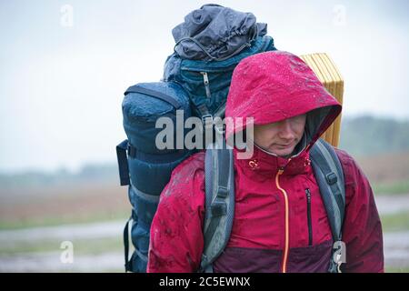 Guy Tourist Wanderer mit Rucksack und Reisematte, in einer roten Jacke steht im Regen Stockfoto